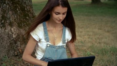 a laptop içinde oturan uzun saçlı güzel Bayan bir ağacın yanında görünüyor