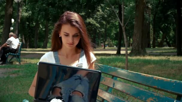 Mooie jonge brunette met zwarte laptop zit op een bankje — Stockvideo