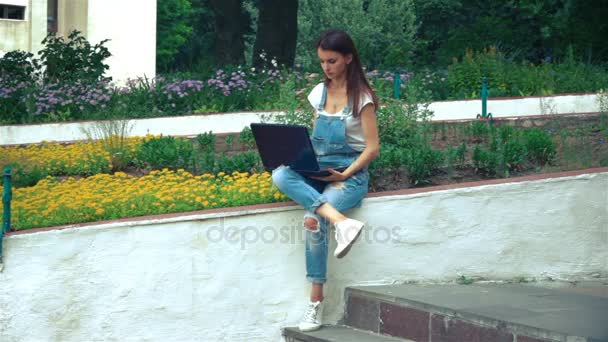 Young girl sits near the flowerbeds with colors and prints on a laptop — Stock Video