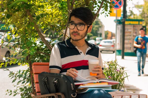 Snygg kille i glasögon håller ett glas kaffe och sitter på en bänk — Stockfoto