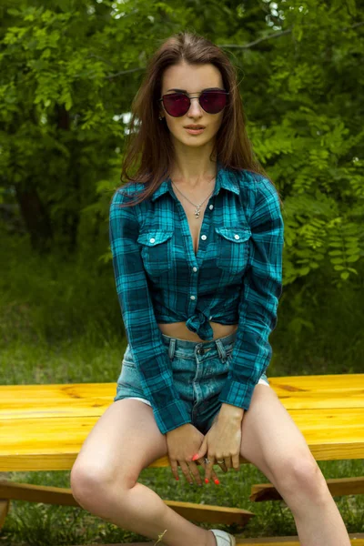 Молодая стройная сексуальная девушка в рубашке и очках, сидящая на скамейке — стоковое фото