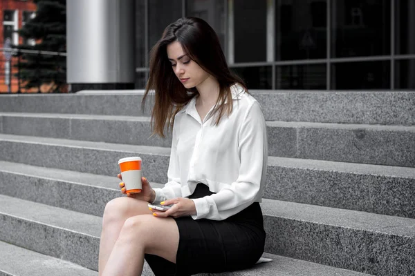 Linda chica de negocios sentado en las escaleras mantiene el café y mira fijamente al teléfono — Foto de Stock