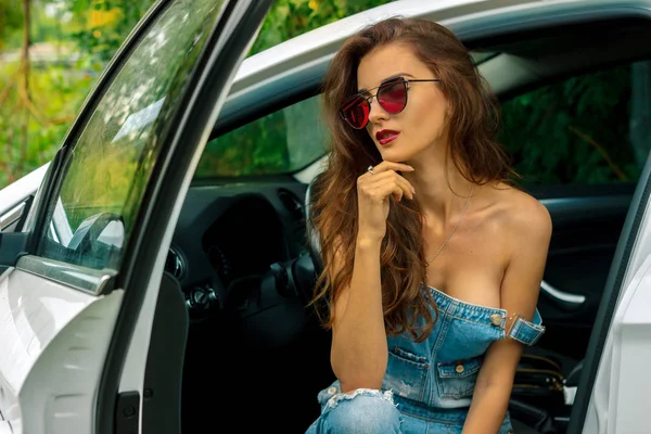 Prachtige jonge brunette meisje met sexy lichaam in spijkerbroek algemene zit in een auto — Stockfoto