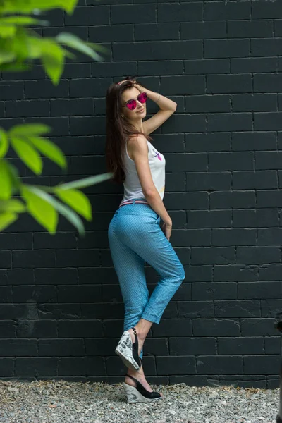 Hermosa joven morena con cuerpo delgado y gafas brillantes se encuentra cerca de una pared — Foto de Stock