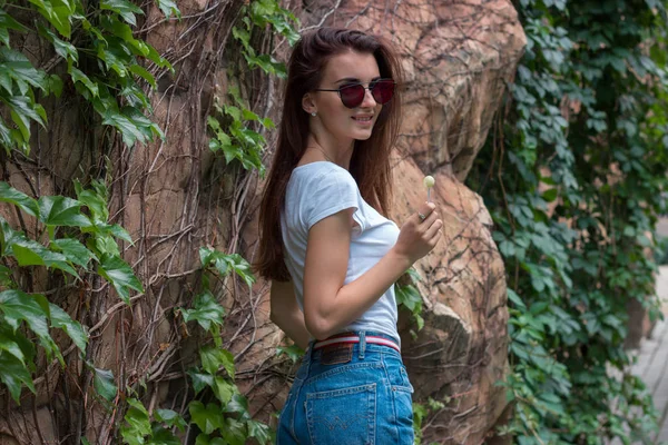 Atractiva morena joven en gafas de espejo se encuentra en un parque cerca de una pared con verdes —  Fotos de Stock