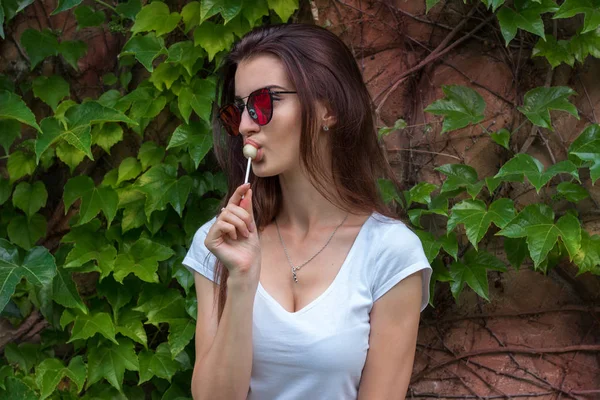 Sexy young brunette girl in sunglasses licks a lollipop — Stock Photo, Image