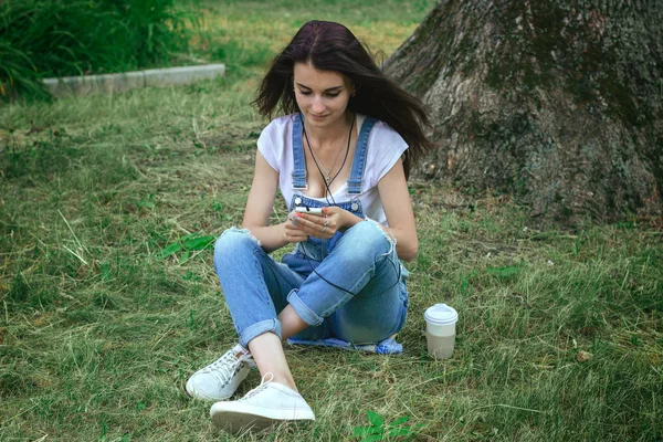 Jeune charmante brune s'assoit dans l'herbe écoutant de la musique et regardant le téléphone — Photo