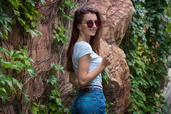 Ελκυστική νεαρή μελαχρινή με είδωλο γυαλιά βρίσκεται σε ένα πάρκο κοντά σε ένα τοίχο με χόρτα — Φωτογραφία Αρχείου
