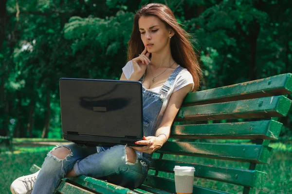 Милая молодая девушка сидит на скамейке в парке с черным ноутбуком — стоковое фото