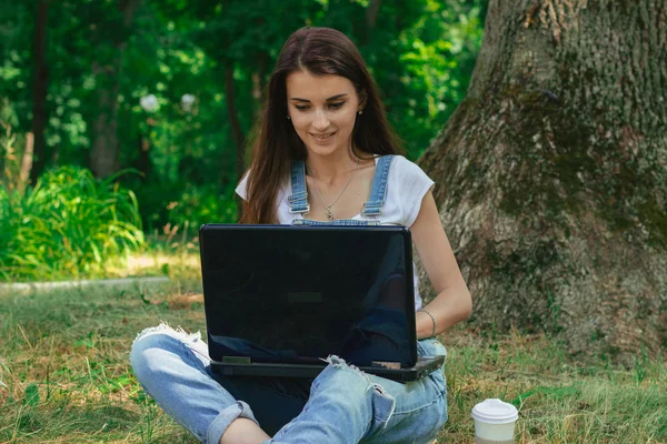 Молодая улыбающаяся женщина сидит с большим черным ноутбуком в парке — стоковое фото