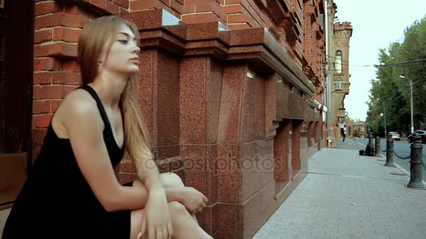Очаровательная одинокая блондинка сидит на улице в черном платье — стоковое видео