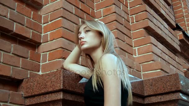 Mooie jonge blonde staat in de buurt van het huis van baksteen en ziet er weg — Stockvideo