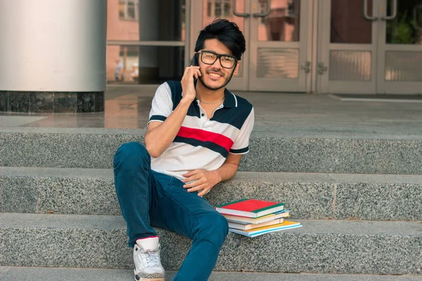 Uśmiechnięty młody facet siedzi na ulicy z książek i rozmawia przez telefon — Zdjęcie stockowe