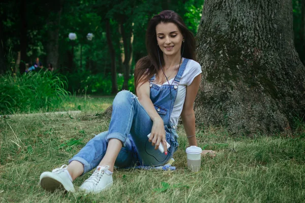 Feliz joven sentada cerca de un árbol en la hierba y escuchando música — Foto de Stock