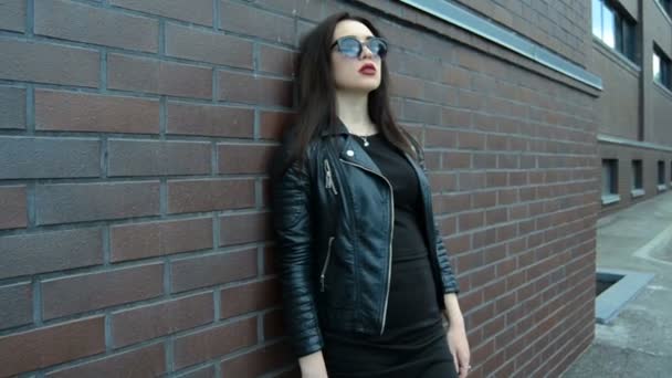 Attraktive junge Brünette in schwarzer Kleidung und Sonnenbrille steht an einer Wand und posiert mit einer Kamera — Stockvideo