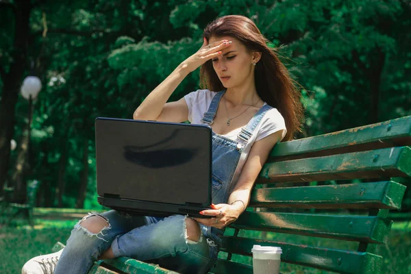 Una joven está sentada en un banco en el parque con un portátil — Foto de Stock
