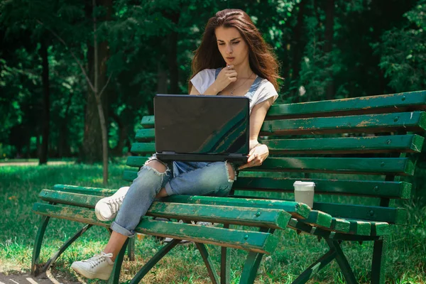 Morena delgada se sienta en un banco en el parque y las impresiones en un ordenador portátil — Foto de Stock