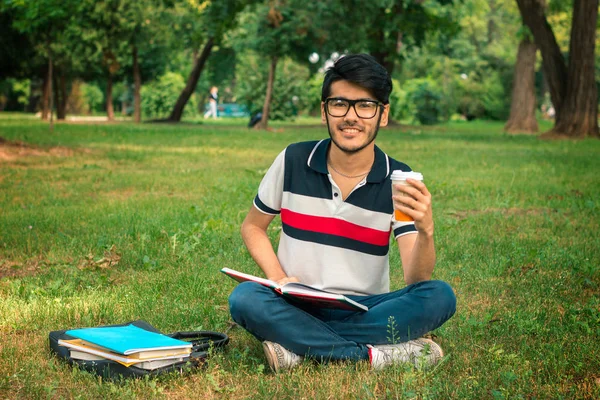 Uśmiechający się facet siedzi na trawie, picia kawy i czytanie książek — Zdjęcie stockowe