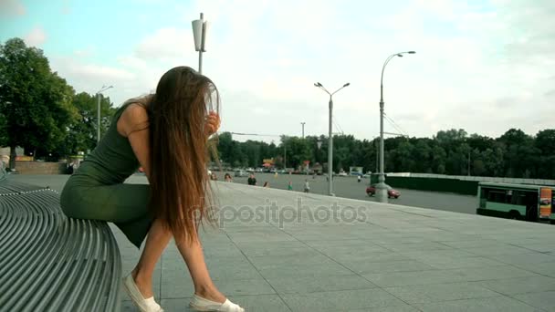 Красивая молодая леди с длинными волосами, сидящая на скамейке и наклоняющая голову — стоковое видео