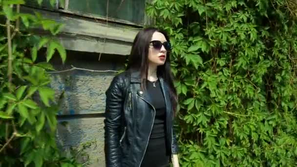 Молодая красивая брюнетка с сексуальными губами в черном стоит рядом с зелеными листьями и ходит по городу позируя на камеру — стоковое видео
