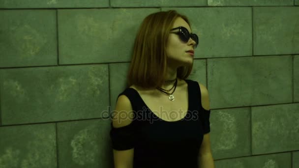 メガネと黒の孤独な女性 t シャツは、歩行者専用地下道の壁近くに立っています。 — ストック動画