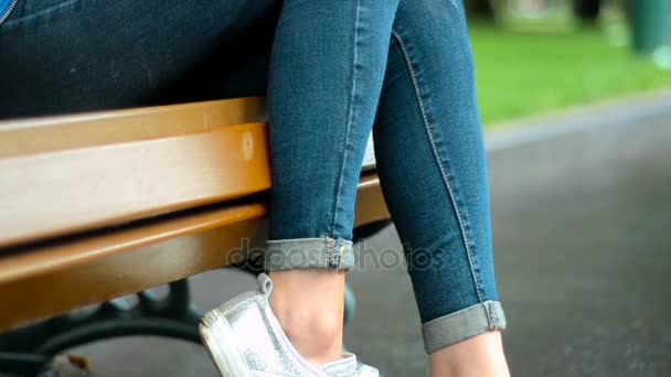 Красива дівчина в білих кросівках і джинсах сидить на лавці — стокове відео