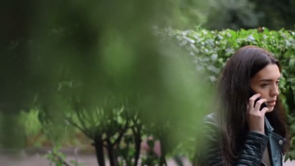 Красивая брюнетка гуляет по парку между деревьями и разговаривает по телефону — стоковое видео
