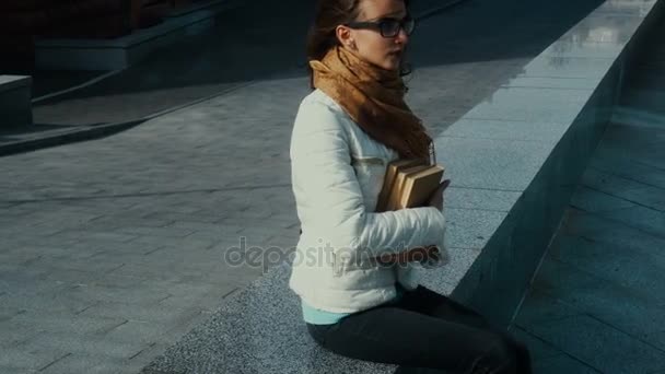 Encantadora joven morena en gafas con libros en las manos se sienta en la calle — Vídeo de stock