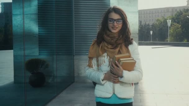 Beautiful brunette woman with books in hands walks in the street and smiles — Stock Video