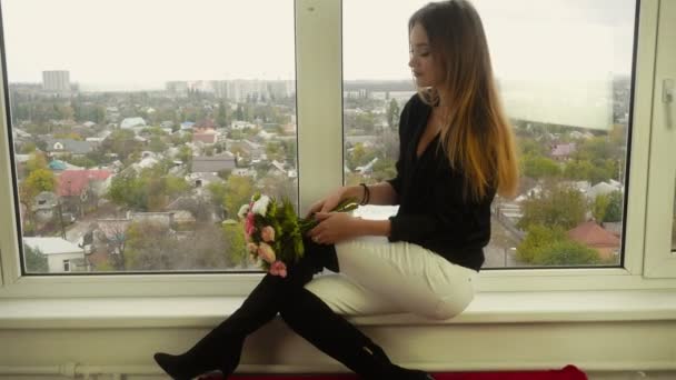 Atrakcyjna dziewczyna siedzi na parapet okna, uśmiechając się i wąchania kwiatów — Wideo stockowe