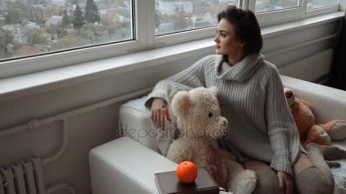 esmer güzel bir oyuncak ayı ile kanepede oturur ve pencereden dışarı vasıl ev görünüyor