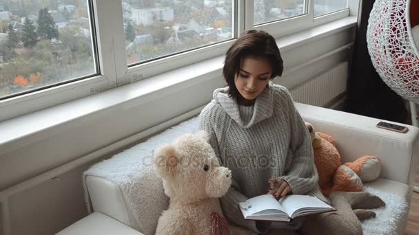 朝は家で本と窓際のソファの上に座っているかわいいブルネット — ストック動画