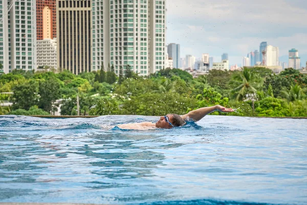 A man swims a butterfly in a pool on the roof of a luxury hotel. View of the city of Manila from the pool of the luxury five-star Discovery Primea hotel. Sunny weather. Skyscrapers on the background.