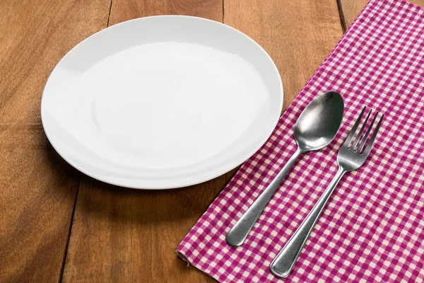 Spoon and fork on tablecloth with white plate on wooden table — Stock Photo, Image
