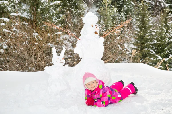Joyeux beau enfant fille plaçant avec un bonhomme de neige sur la marche hivernale enneigée. Bâtiment enfant bonhomme de neige à Noël . — Photo