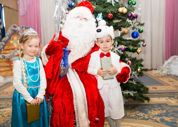 Duas crianças vestidas em ternos de carnaval com Papai Noel perto de abeto de Natal em férias de crianças de Ano Novo com presentes — Fotografia de Stock