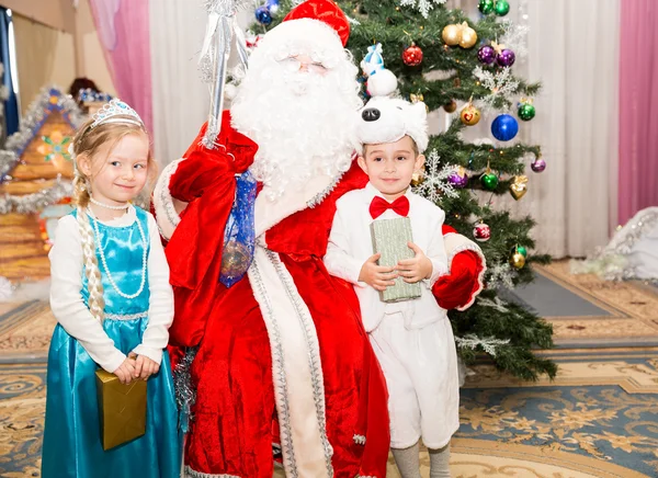 Duas crianças vestidas em ternos de carnaval com Papai Noel perto de abeto de Natal em férias de crianças de Ano Novo com presentes — Fotografia de Stock