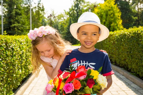 Black african american boy kid gives flowers to girl child on birthday. Little adorable children in park. Childhood and love. — Stock Photo, Image