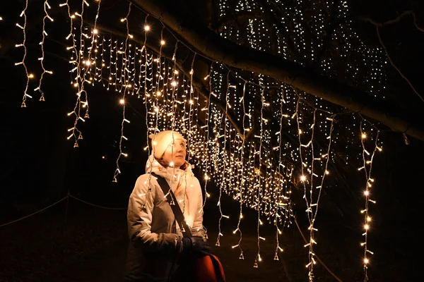 Shiny glowing white lights in night darkness and young woman among hanging string lights  garlands. Cheerful woman and shiny blurry Christmas lights on black background. Winter holiday mood. Night scene