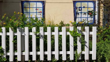 İki sahte pencereli sarı duvarlı evin önündeki beyaz boyalı ahşap çit. Beyaz çitlerin arkasında büyüyen renkli çiçeklerden oluşan çiçek tarhı. İlkbaharın huzurlu manzarası