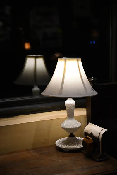 台灯典雅的白色古董台灯特写在黑暗模糊的反射上 灯的轮廓模糊了玻璃窗中的反射光 古老的白色灯罩在黑暗中闪闪发光 — 图库照片