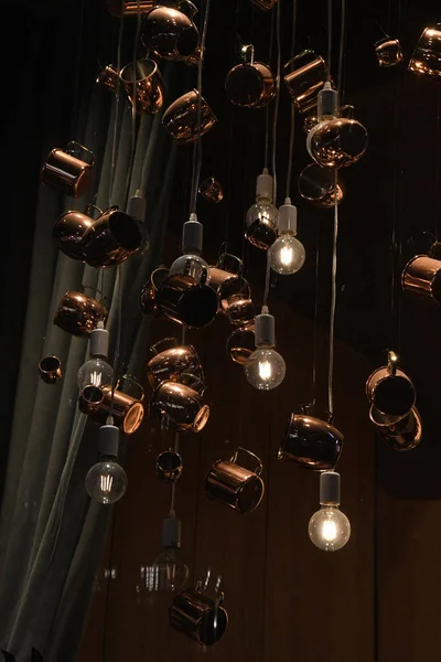 Retro style pendant lights made from glossy copper mugs and shiny light bulbs hanging on wire strings. Unusual modern lighting equipment.