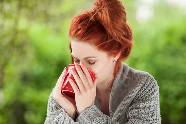 美丽的年轻女子 红色的头发坐在花园里 喝咖啡在一个红色的咖啡杯 — 图库照片