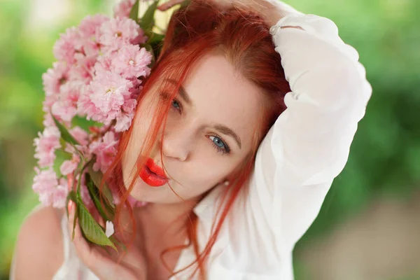 Beautiful young woman with red hair having fun standing in the garden with cherry blossom branch and perfect make up