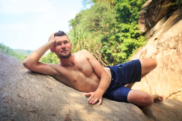 closeup Avrupa tabaklanmış sakallı adam şort kayada bared gövde yalanlarla mesafe içine tropik bitkiler tarafından görünüyor.