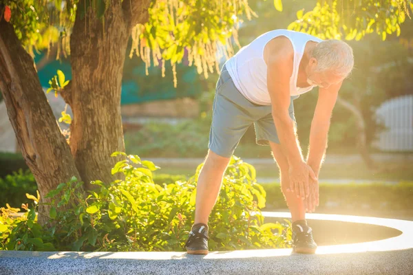 bearded grey old man in vest shorts does morning exercises bends body to foot on stone barrier against sun backlight