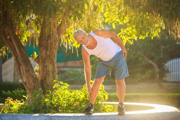 bearded grey old man does morning exercises bends body to foot on stone barrier against sun backlight