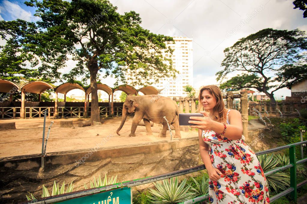 blond girl in flowery frock makes selfie against elephant in resort city zoo
