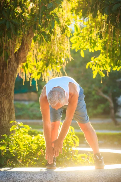 old man in vest shorts does morning exercises bends body to foot on marble barrier against sun