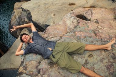 Güneş ışığı gülümsüyor altında dağ nehir taşları üzerinde gömlek ve şort yalan Avrupa kıvırcık adam V gösterir